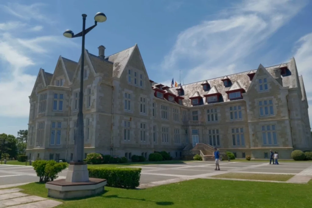 Die weitläufige, schlossähnliche Rückseite des Palacio de la Magdalena in Santander mit einer großen Freifläche davor. Eine markante, dunkle Straßenlaterne steht im linken Vordergrund unter leicht bewölktem Himmel.
