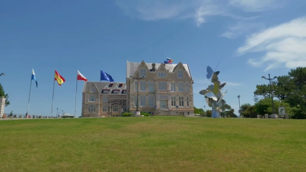 Der Palacio de la Magdalena von einer grünen Wiese aus fotografiert. Links wehen Flaggen im Wind, rechts steht eine moderne, bunte Skulptur aus großen Schmetterlingen.