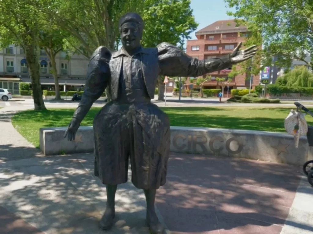 Eine lebensgroße Bronzestatue des eleganten Weißclowns Manolo Tonetti im Parque de Mesones in Santander. Die Figur steht mit weit geöffneten Armen auf einem gepflasterten Platz vor einer niedrigen Mauer mit der Aufschrift „CIRCO“. Im Hintergrund befinden sich grüne Parkbäume und Wohngebäude.