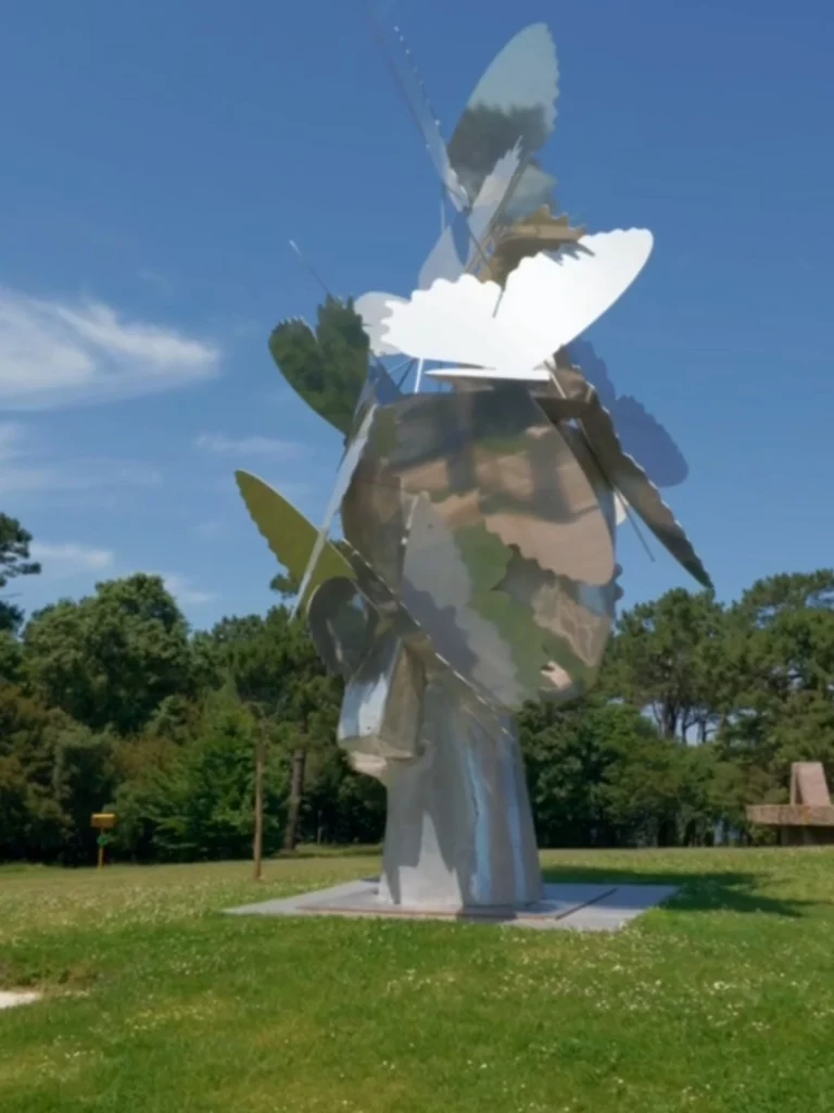 Eine moderne, große Skulptur aus spiegelndem Metall in Form eines menschlichen Kopfes, auf dem zahlreiche silberne Schmetterlinge sitzen, platziert auf einer grünen Wiese vor Bäumen.