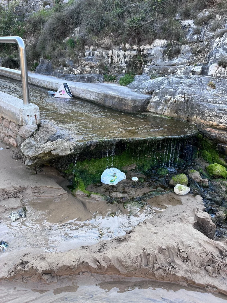 Ein kleiner Bachlauf fließt über Felsen in den Sandstrand, leider liegt Plastikmüll im Wasser.