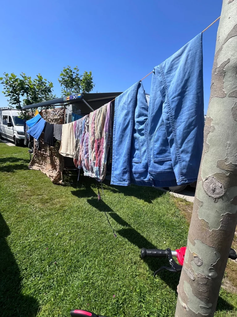 Jeans und bunte Kleidung hängen zum Trocknen an einer langen Wäscheleine auf einem sonnigen Campingplatz.