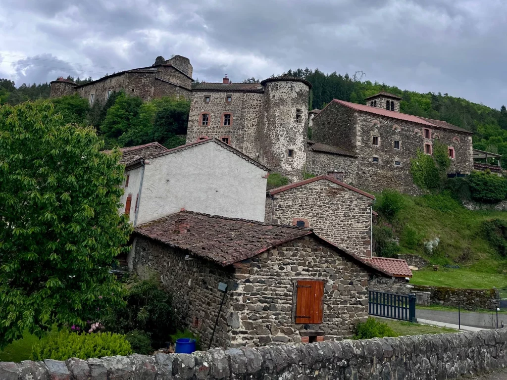 Alte, rustikale Steinhäuser und eine historische Burganlage mit Rundturm an einem grünen Hang in Frankreich.