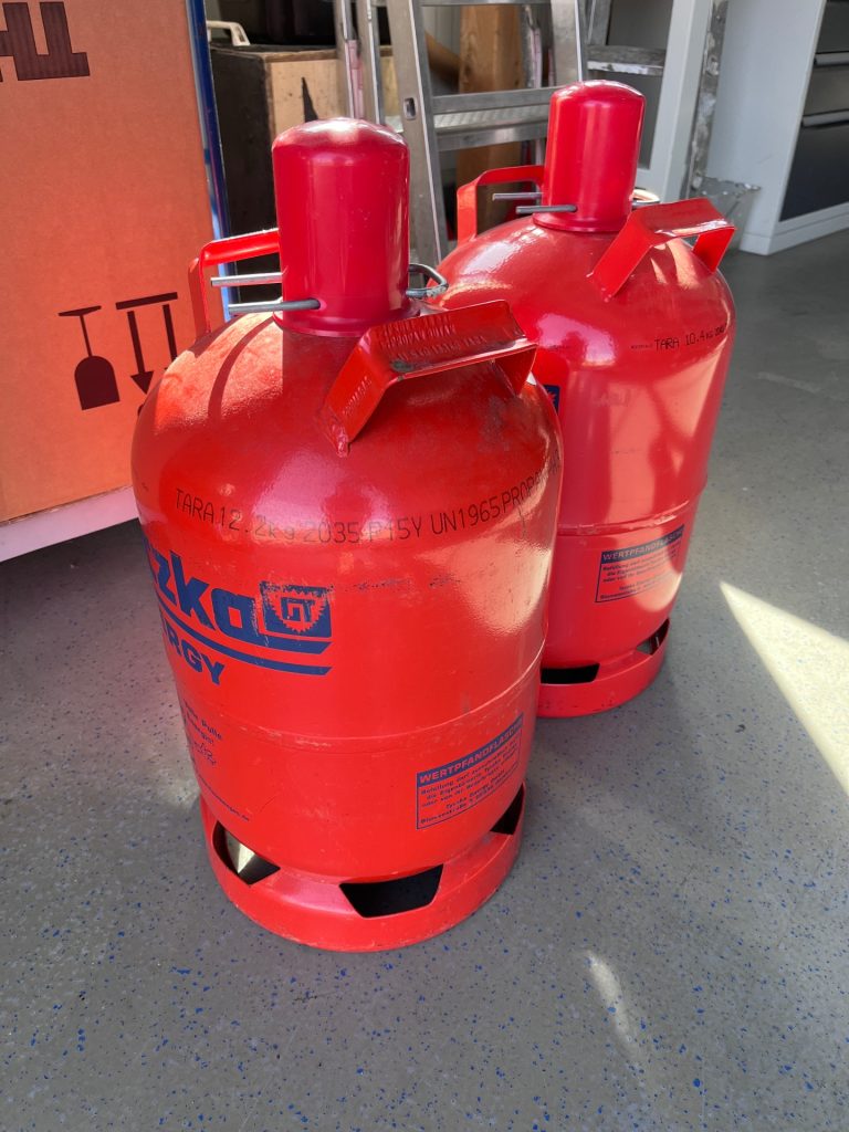 Zwei rote 11-kg-Gasflaschen stehen nebeneinander am Boden vor einer Werkstatt.